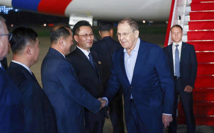 Several officials in dark business suits greet each other with handshakes. The scene takes place near a large aircraft with visible red and white coloring. 