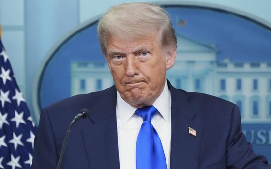 Donald Trump, wearing a dark suit with a U.S. flag pin, stands at a microphone in front of a large White House seal on the wall behind him.