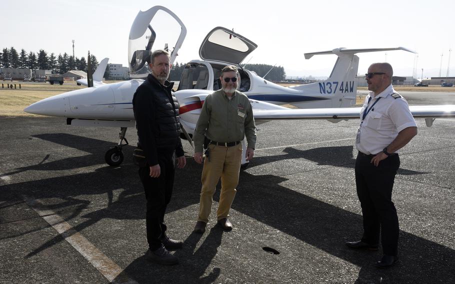 Three men stand on a runway in front of a small aircraft with its cockpit hatch and rear doors open.
