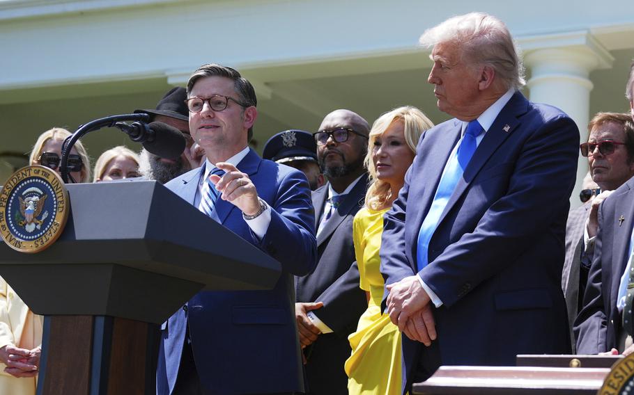 Mike Johnson stands at a podium and speaks into a microphone as Donald Trump stands to the side.