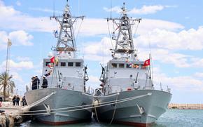 Two gray military patrol boats are docked side by side at a pier, adorned with colorful signal flags and the red Tunisian national flag. Several naval personnel stand on the deck of the left vessel, and a group of people is visible onshore. 