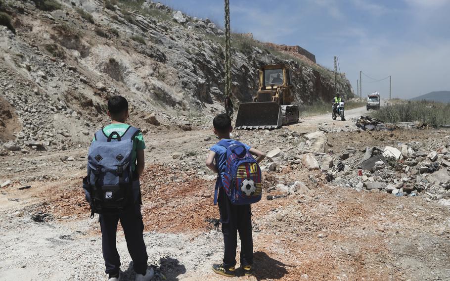 Two children with backpacks look on as a bulldozer shovels dirt in a hilly/mountainous area.