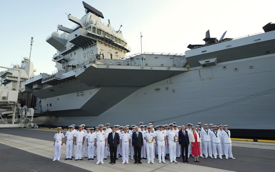 A group of British sailors at a dock next to an aircraft carrier.