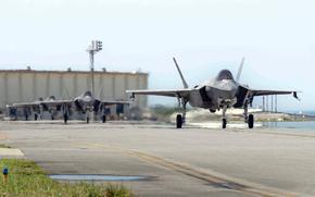 A row of F-35 fighter jets taxis down a runway on a bright day, with the lead jet front and center and a few more lined up behind it. Heat waves rise from the pavement, and hangars are visible in the background.