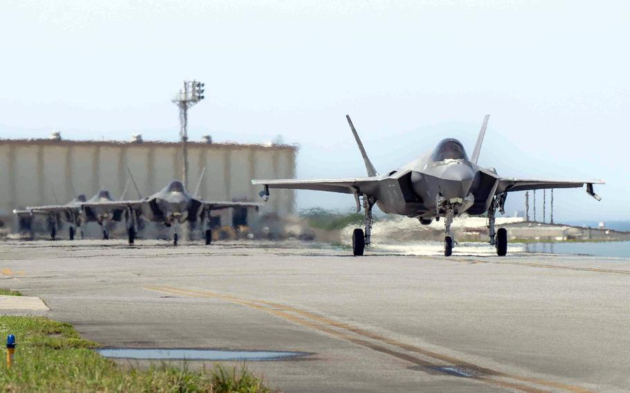A row of F-35 fighter jets taxis down a runway on a bright day, with the lead jet front and center and a few more lined up behind it. Heat waves rise from the pavement, and hangars are visible in the background.