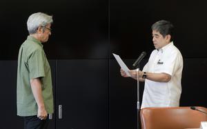 Two men are standing in a formal setting. The man on the right, wearing a white shirt and a smartwatch, is reading from a piece of paper into a microphone. The man on the left, wearing a green shirt, listens attentively. 