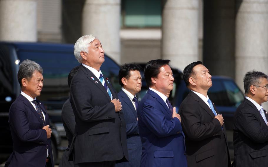 A group of formally dressed officials in dark business suits stand in a line with their hands placed over their hearts. 