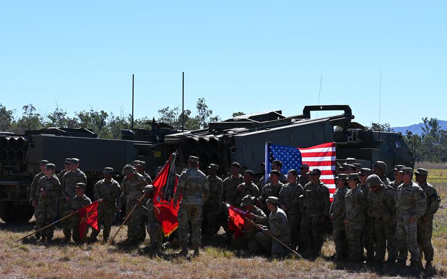 Soldiers stand in front of an American flag flanked by HIMARS launchers on a sunny day.