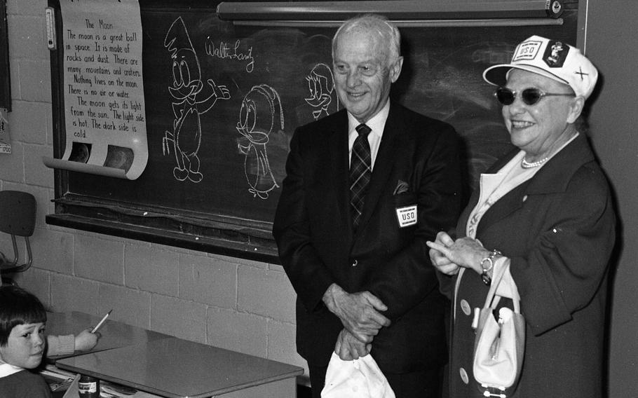 An elderly couple smiling near a blackboard with cartoon characters drawn on it.