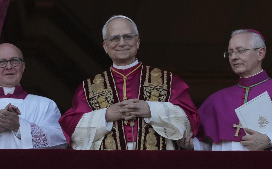 Pope Leo XIV seen from the waist up, smiling and clasping his hands as he stands at a basilica.