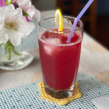 Close-up view of a glass with a red-pink drink, lemon wedge on the rim and purple straw sitting on a yellow coaster atop a blue checkered placemat. A small vase with white flowers is behind the glass.