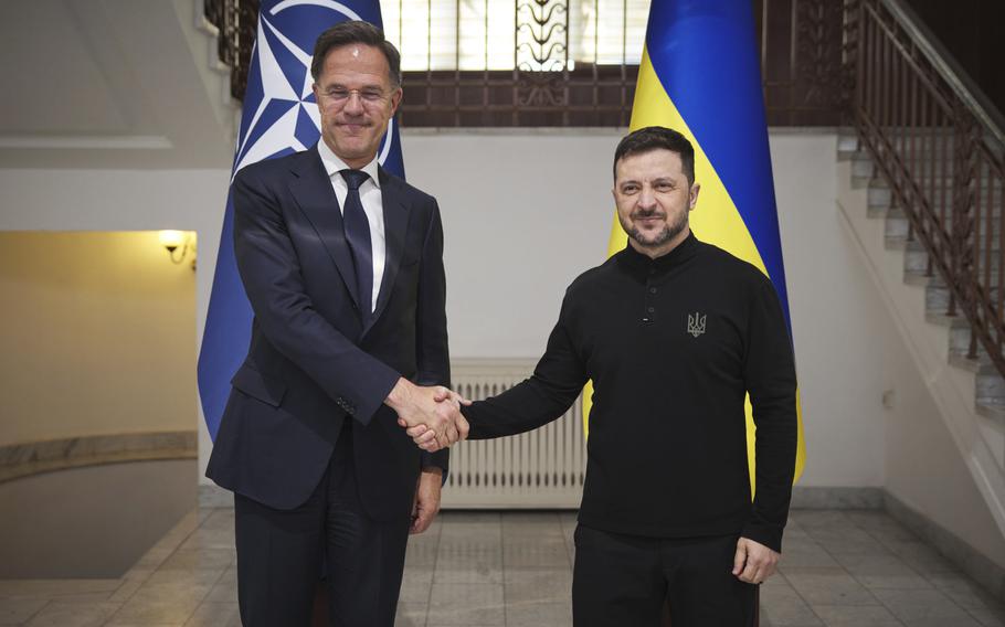 Volodymyr Zelenskyy and Mark Rutte shake hands while posing in front of Ukraine and NATO flags.