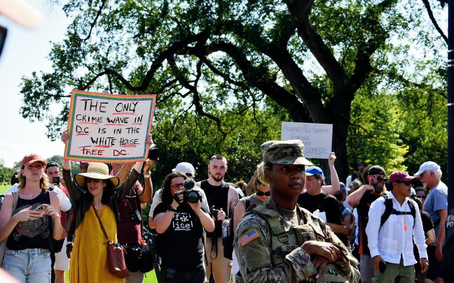 A lone National Guard soldier stands in front of a crowd of protesters. One protester holds a sign that reads: “The only crime wave in DC is in the White House. Free DC.”