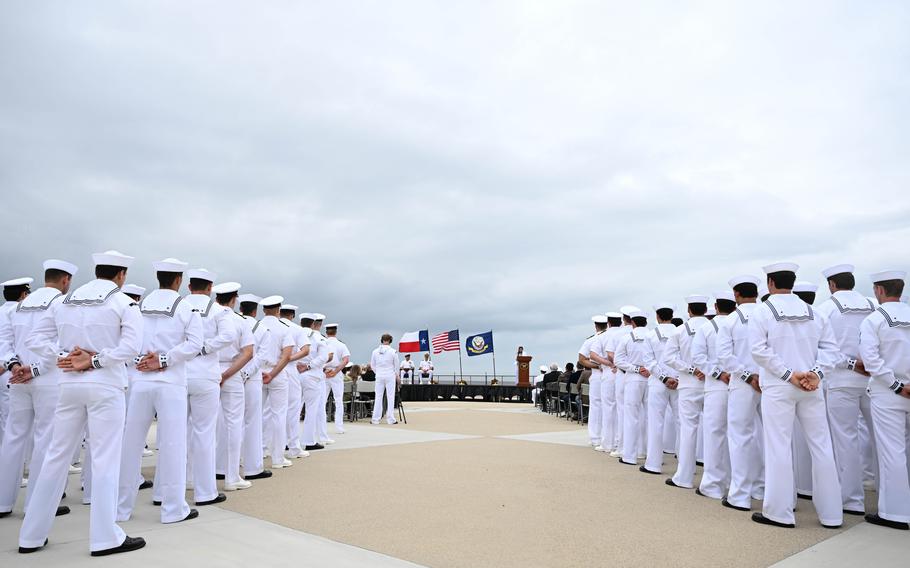 Navy Special Warfare operators in white uniforms stand in formation on either side of the photo, while the center of the image is an open path leading up to a stage with a speaker at a podium.