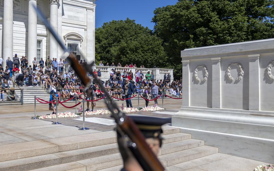 A member of the honor guard in the foreground with a rifle on his shoulder marches past the tomb. A crowd of people gathers in the background.