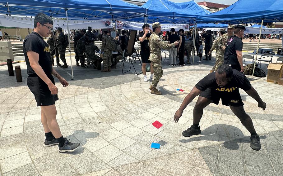 Soldiers in black shorts and t-shirts play a game with red and blue cards on a concrete patio with other soldiers and blue canopies in the background.
