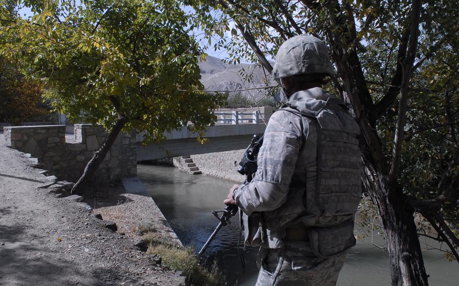 A soldier, seen from behind, patrolling by a river in a mountainous area.