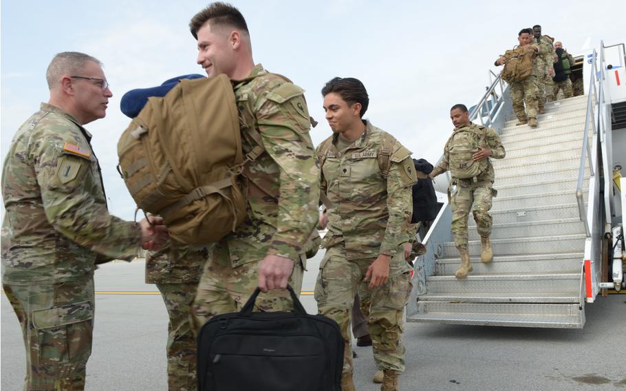 A line of soldiers walks off the steps of a plane, with those closest to the camera shaking the hand of one of their leaders.