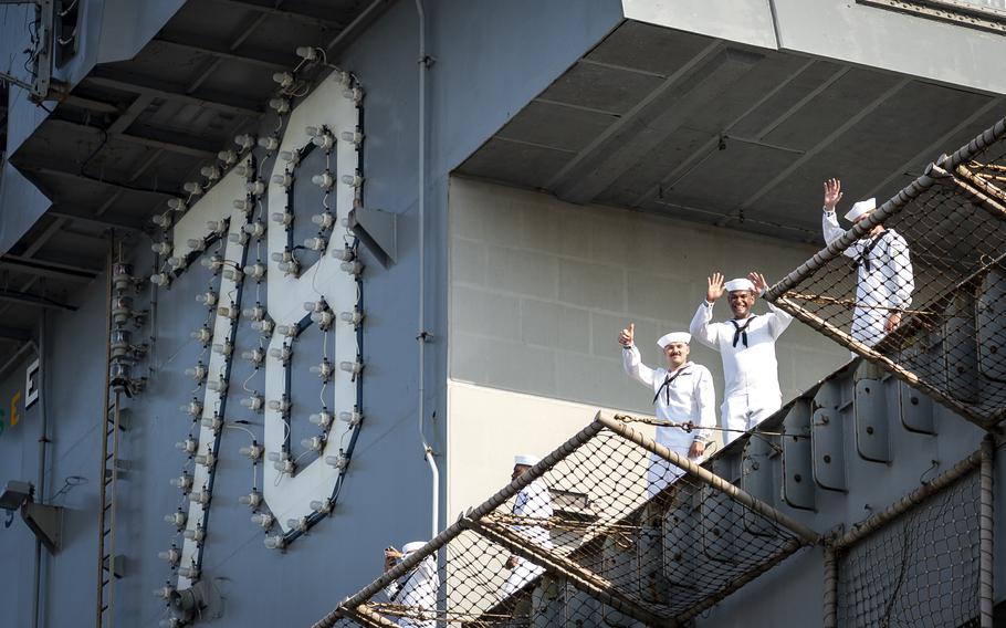 Three sailors in white uniforms and hats, seen from below, wave while standing near the rail aboard an aircraft carrier.