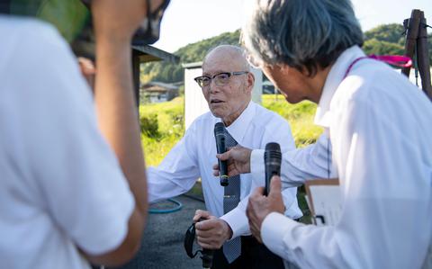 An elderly Japanese man wearing a white shirt, green tie and dark-rimmed glasses stands with the assistance on a cane, while another man to the right holds a microphone in front of him and a person to the left records with a video camera.