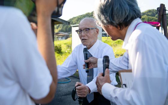 An elderly Japanese man wearing a white shirt, green tie and dark-rimmed glasses stands with the assistance on a cane, while another man to the right holds a microphone in front of him and a person to the left records with a video camera.