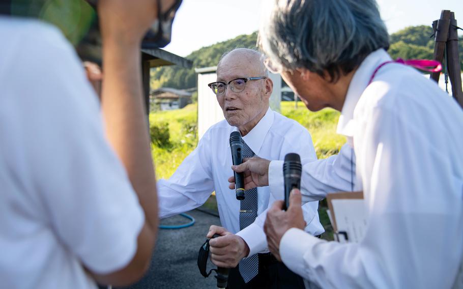 An elderly Japanese man wearing a white shirt, green tie and dark-rimmed glasses stands with the assistance on a cane, while another man to the right holds a microphone in front of him and a person to the left records with a video camera.