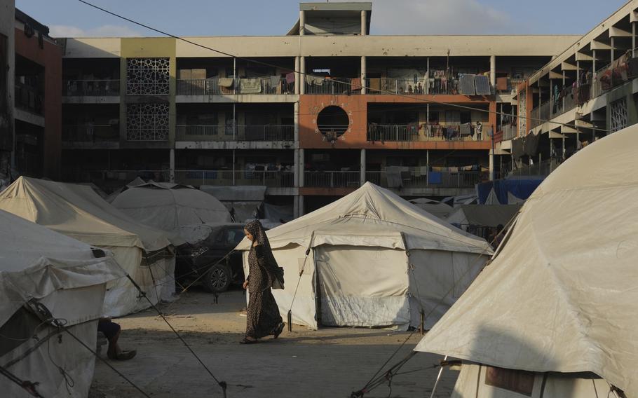 A woman wearing a black dress and head scarf walks among white canvas tents in a courtyard with a multi-story building in the background.