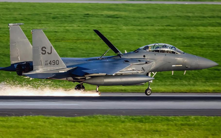 A gray F-15E Strike Eagle fighter jet lands on a runway, sending sparks flying froma missing right back wheel. Green grass can be seen in the background.