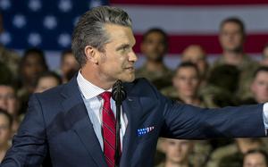 Pete Hegseth looks to the side and points with his arm outstretched while standing at a microphone in front of military service members in camouflage uniforms and a large U.S. flag.