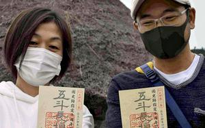Two Japanese people wearing facemasks hold up small planks of wood with red stamps and Japanese writing.