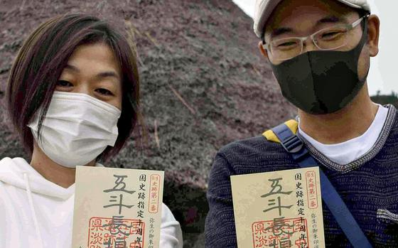 Two Japanese people wearing facemasks hold up small planks of wood with red stamps and Japanese writing.