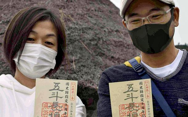 Two Japanese people wearing facemasks hold up small planks of wood with red stamps and Japanese writing.