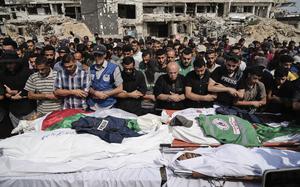 A crowd of Palestinian mourners bows their heads and pray next two several bodies covered by white shrouds and with a blue vest reading “PRESS” lying on one.