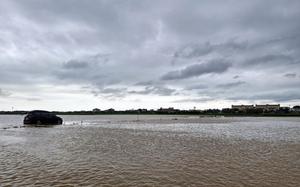 A dark-colored SUV sits partially submerged in muddy floodwater under a cloudy sky. 