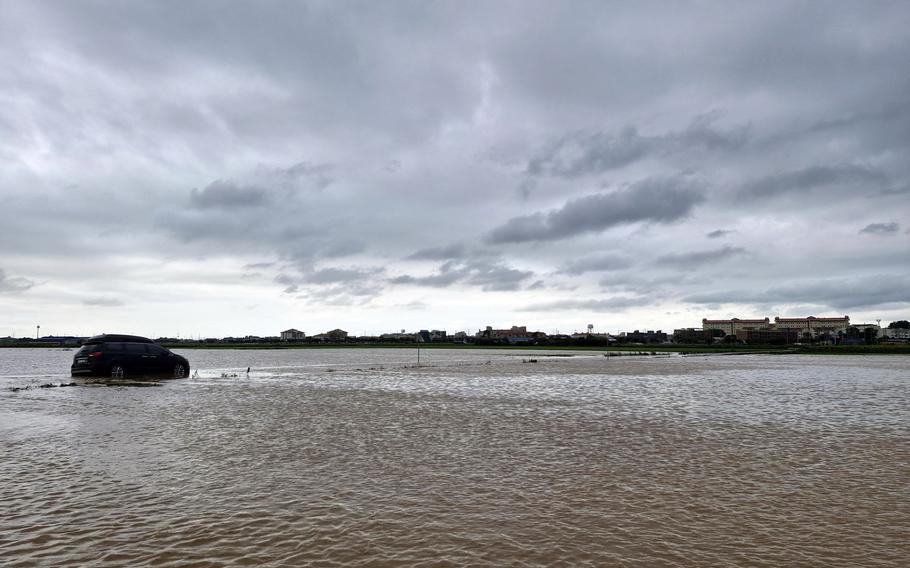 A dark-colored SUV sits partially submerged in muddy floodwater under a cloudy sky. 