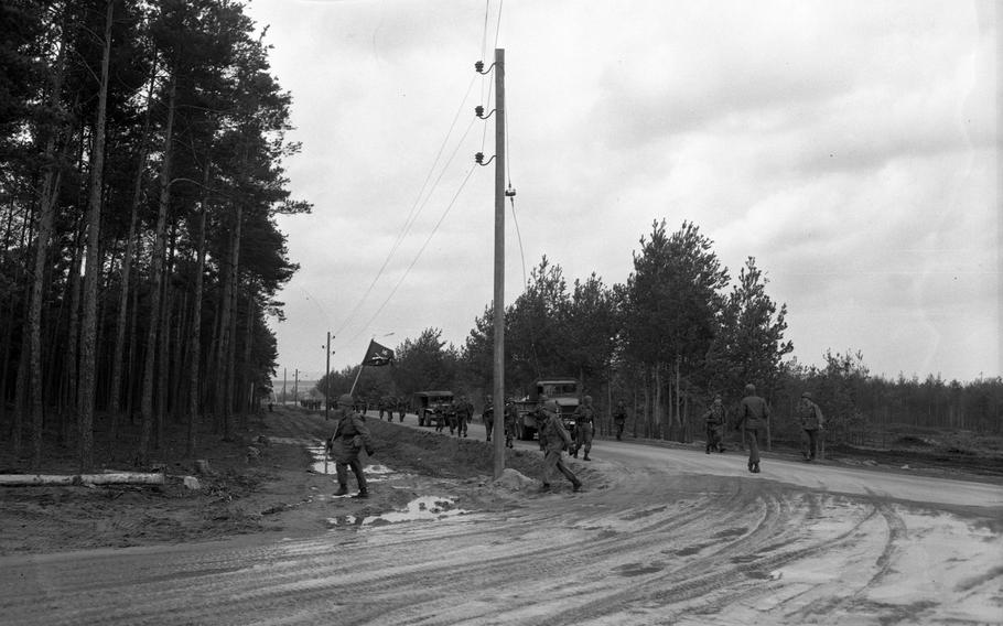 A black and white image of soldiers and military vehicles passing through the corner of a muddy road.