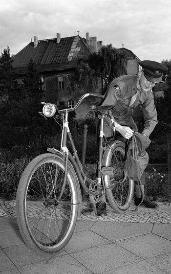 A vertical, black and white photo of a service member adjusting a bad at the back of his bicycle.