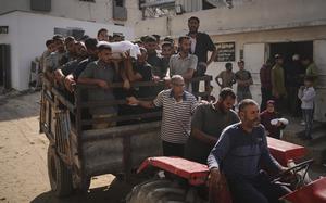 A group of Palestinian men carry a shroud-covered body while standing in a wagon being pulled by a man on a tractor.
