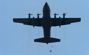A military transport aircraft silhouetted against a clear blue sky, viewed from below, with four propeller engines and a cargo or personnel package being dropped by parachute beneath the aircraft.