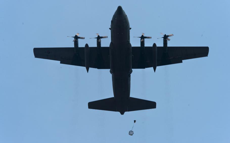 A military transport aircraft silhouetted against a clear blue sky, viewed from below, with four propeller engines and a cargo or personnel package being dropped by parachute beneath the aircraft.