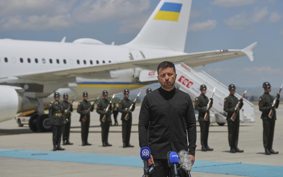 Volodymyr Zelenskyy speaks in front of several microphones on an airport’s tarmac, while an military personnel and an airplane stands behind him.