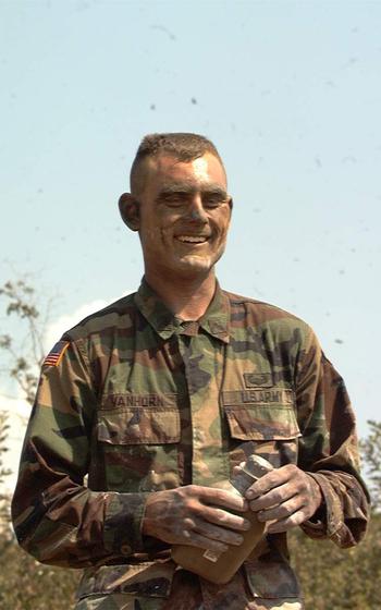 A soldier with a dirtied face smiles while holding a canteen or bottle.