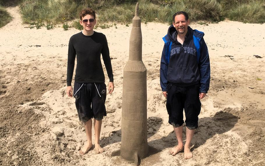 A sand sculpture of the Saturn V rocket in between two 6-foot-tall men. 