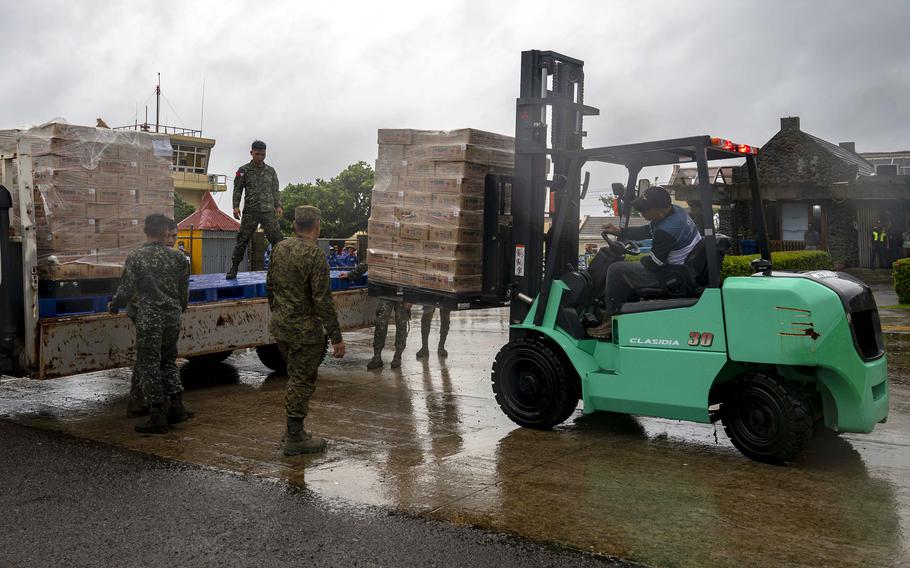 A man operating a green forklift removes a pallet of cardboard boxes from the back of a flatbed truck as military service members in camouflage uniforms observe.