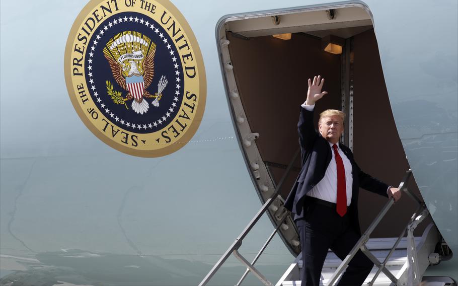 Donald Trump, wearing a dark suit and red tie, waves while standing at the top of a staircase next to an airplane with the seal of the President of the United States.