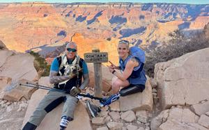 A man and a woman, each with a prosthetic leg and wearing hiking gear, sit on rocks and pose with a small wooden sign reading “Ooh Aah Point” between and a Grand Canyon vista behind them.
