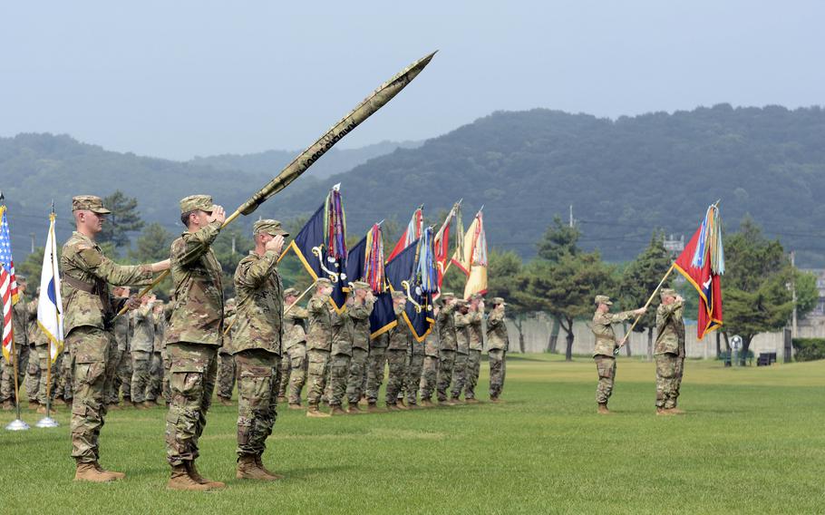 Two soldiers in camouflage uniforms stand at attention and salute on a grassy field while a line of soldiers in camouflage uniforms stand in the background holding flags.