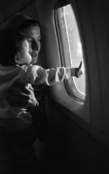 An infant points out the window of a plane while being held by a woman.