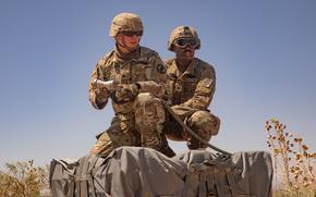 Two soldiers in camouflage combat gear, helmets and sunglasses kneel next to a large gray package of supplies, with blue sky in the background.