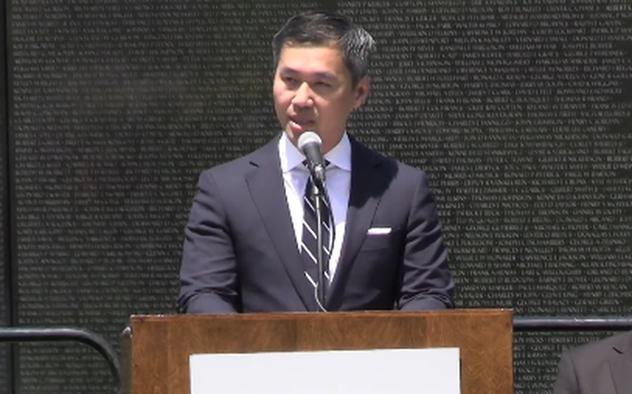 A man in a dark suit stands at a podium and speaks into a microphone in front of a black marble wall engraved with the names of troops killed in war.